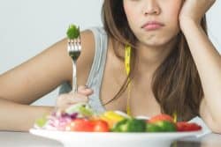 Unhappy woman sitting at a table with a plate of vegetables and broccoli skewered on a fork. She has a tape measure draped over her shoulder.