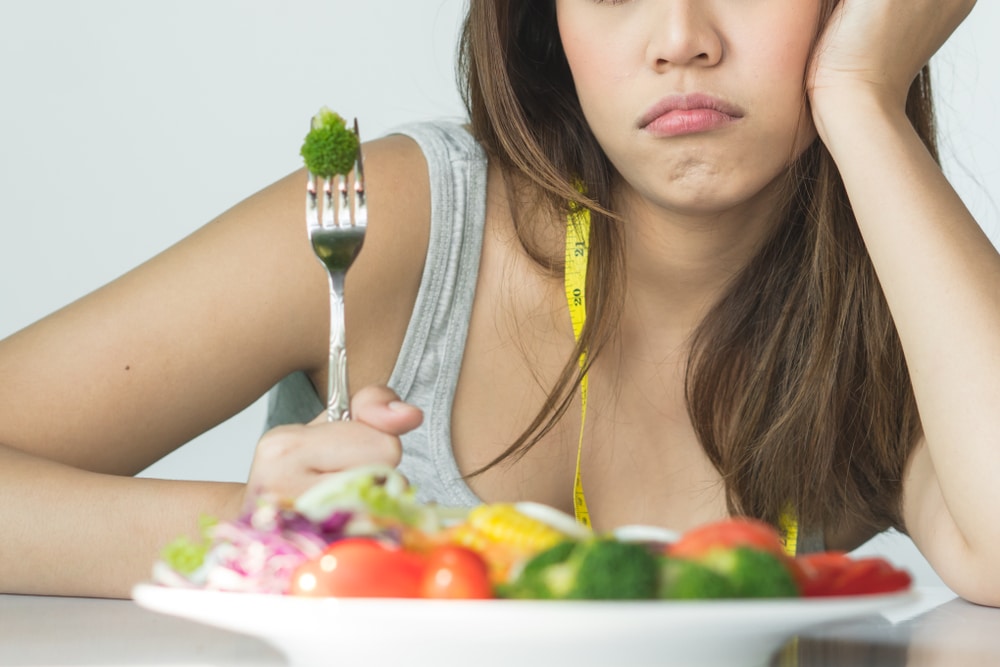 Unhappy woman sitting at a table with a plate of vegetables and broccoli skewered on a fork. She has a tape measure draped over her shoulder.