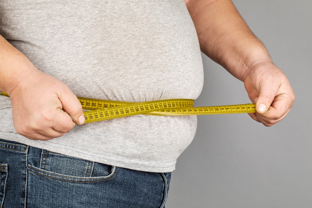 Obese man in grey shirt measures his waist with tape measure