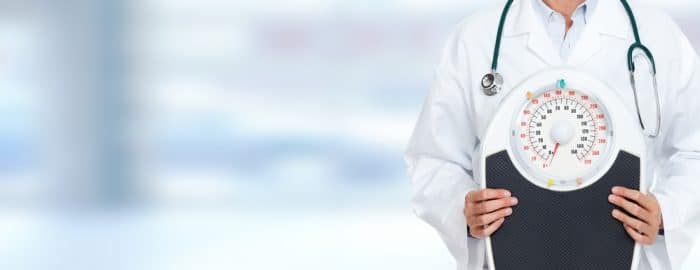 Doctor in lab coat with a stethoscope around his neck holds up an analog scale.