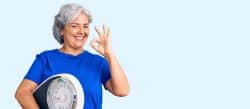 Elderly woman hold scale up to her body with one hand, while making the OK gesture with the other.