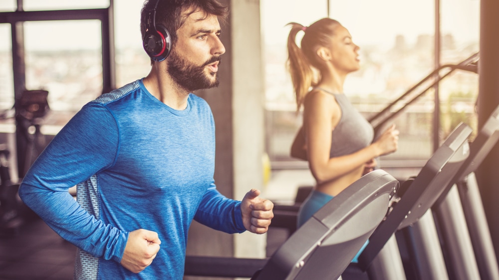 In the foreground a bearded man in a blue shirt is on a treadmill. Behind him is a blonde woman who is also on a treadmill.