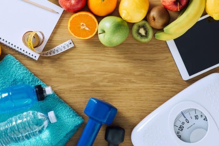 Scale, exercise equipment, fruit tape measure, and journal on a table for workout planning.