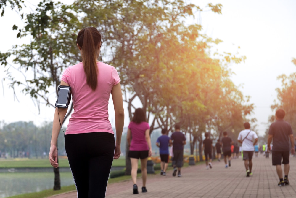 A woman in exercise attire is walking along the edge of a lake in a park with other walkers in the background.