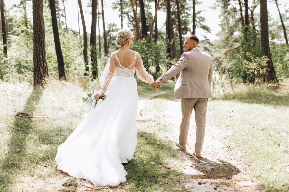A married couple standing together in a forest, holding hands.