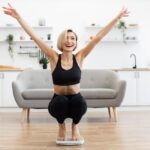 A blonde woman in black athletic wear celebrates excitedly while squatting on a bathroom scale in a bright room