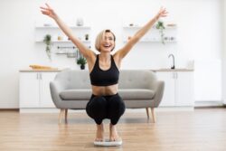 A blonde woman in black athletic wear celebrates excitedly while squatting on a bathroom scale in a bright room