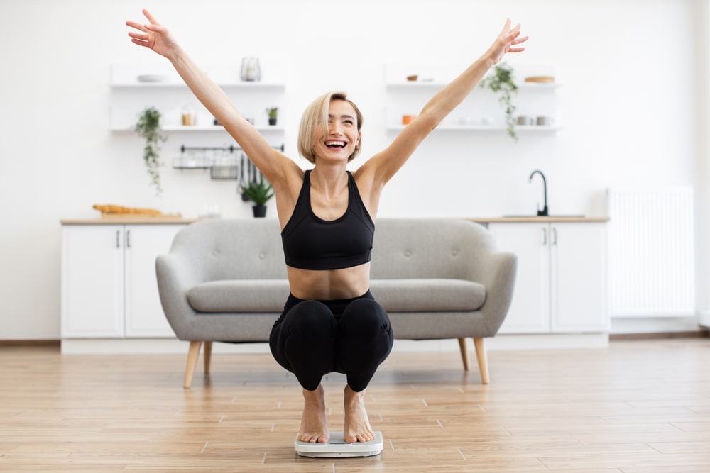 A blonde woman in black athletic wear celebrates excitedly while squatting on a bathroom scale in a bright room